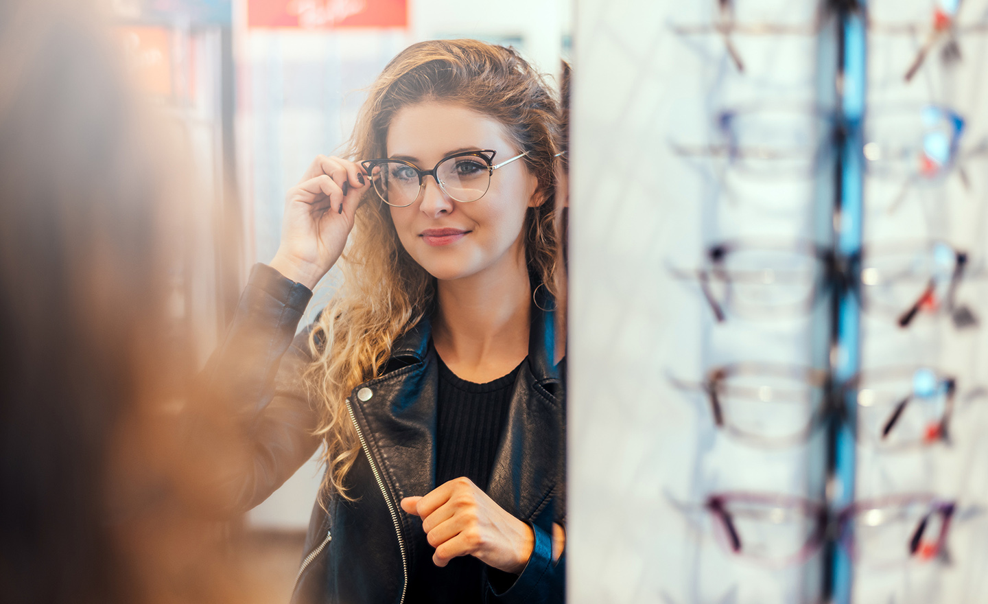 Smiling young woman trying on glasses on mirror in optician.