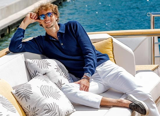 Un homme aux cheveux bouclés, souriant, ajustant ses lunettes de soleil bleues, assis sur un yacht luxueux.