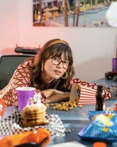 jeune femme portant un frange et des lunettes rectangulaires acoudée sur une table avec des cookies, pop corn et boissons