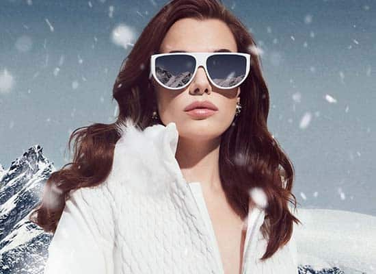 femme avec manteau ski blanc sous la neige portant des lunettes de soleil blanches avec verres solaires bleu miroir sur fond de montagne et neige marque linda farrow-image mise en avant