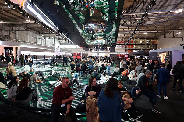 General view of a trade show with visitors resting on wavy floor installations under green and black ceiling decor.