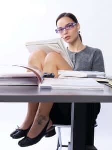 Femme assise à un bureau, les jambes croisées, portant des lunettes de vue bleues et lisant un livre.