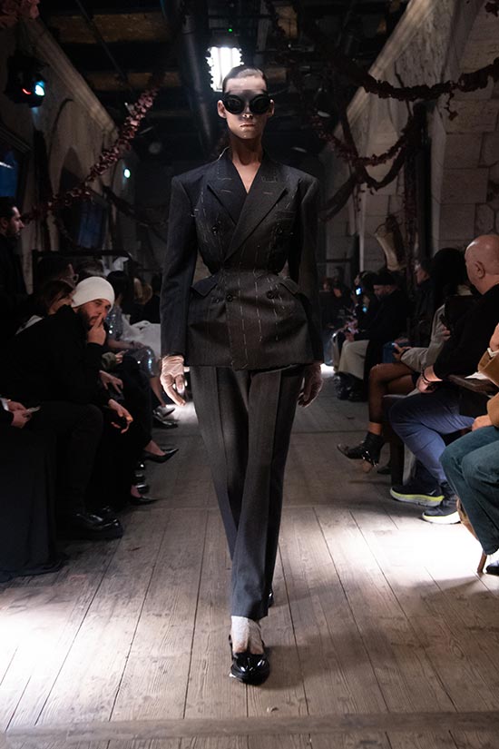 Model walking in gray striped suit, wearing massive black sunglasses, catwalk atmosphere with audience.