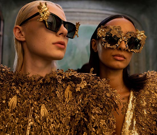Two models wearing extravagant gold embellished sunglasses and luxurious gold clothing.