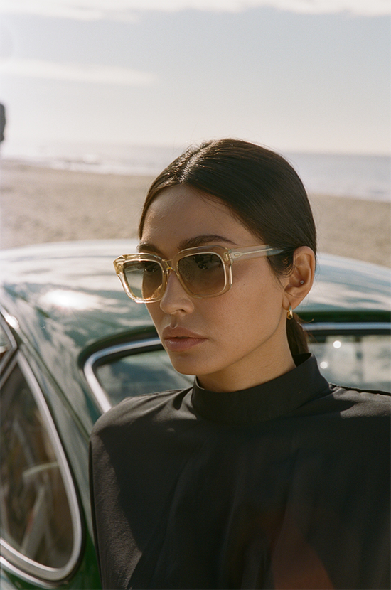 Stylish woman with transparent hexagonal sunglasses leaning against a vintage car on a sunny beach.