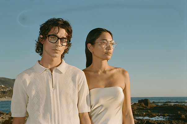 Couple side by side in front of an ocean scene, the man in a white polo shirt, the woman in a white strapless dress, both wearing modern eyeglasses-header article