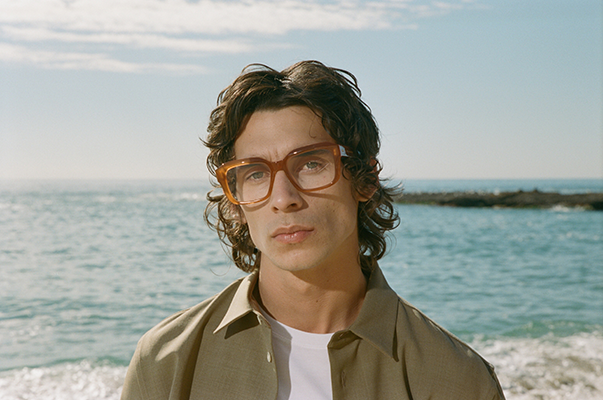 Man with curly hair wearing oversized caramel-colored glasses, looking into the distance on a rocky shore.