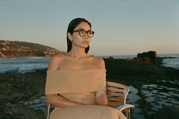 Pensive woman in beige off-the-shoulder dress sitting on a metal chair, wearing clear geometric eyeglasses, at dusk on a beach.
