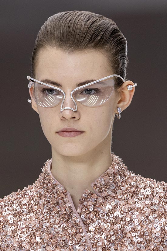 Close-up of a model wearing transparent glasses with rhinestone details, matched with a glittery outfit.