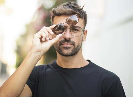 Homme avec une barbe et moustache, soulevant légèrement le clip solaire de ses lunettes rondes, en plein air avec une lumière naturelle douce.