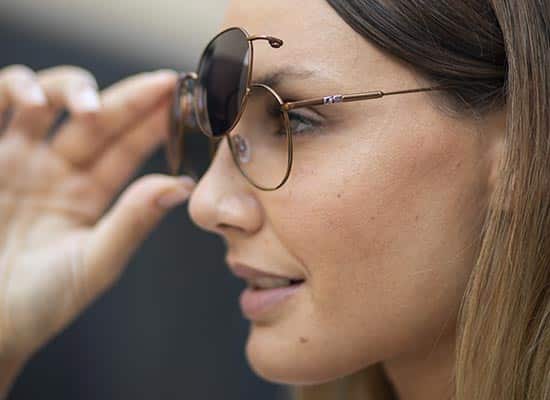 Close-up of a woman's face looking through round sunglasses, highlighting the frame design and tinted lenses.