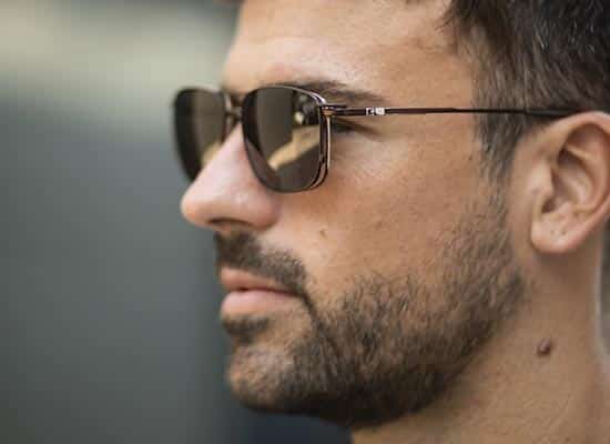 Man with beard and mustache wearing aviator sunglasses with subtle reflection, side profile on neutral background.