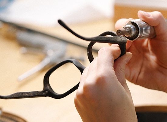 Artisan ajustant méticuleusement une monture de lunettes noire à l'aide d'un outil rotatif.