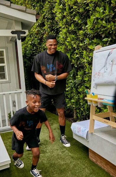 Russel Westbrook et un jeune garçon, tous deux portant des t-shirts noirs et des baskets noires, sourient et jouent dehors près d'un tableau de dessin.