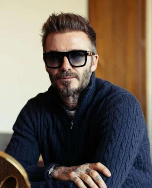 David Beckham portant un pull en maille bleu marine et des lunettes de soleil noires est assis, les bras croisés, avec un léger sourire.