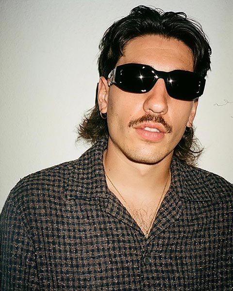 Hector Bellerin avec des cheveux longs, une moustache et des lunettes de soleil noires, portant une chemise à carreaux foncés, pose devant un mur clair.