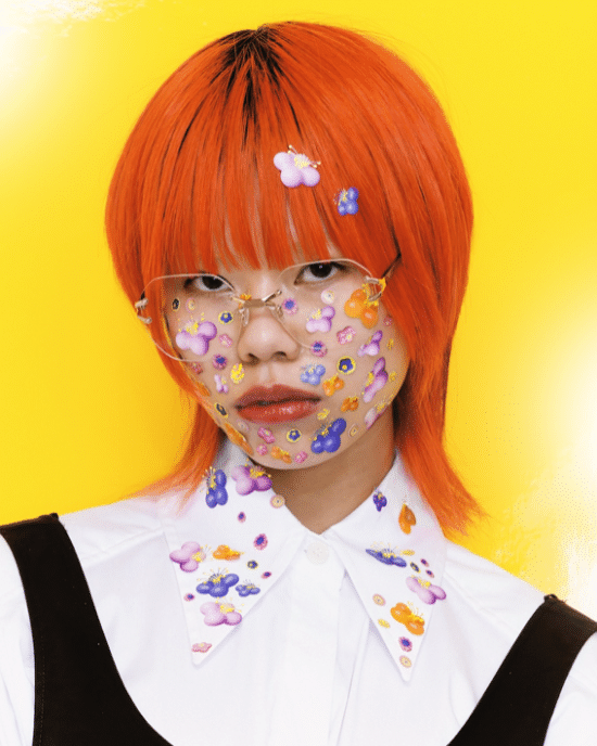 Jeune femme aux cheveux roux vifs portant des lunettes de vue et des autocollants colorés en forme de fleurs sur le visage, posant devant un fond jaune vif.