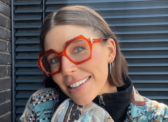 Femme souriante portant des lunettes à monture rouge vif et un manteau coloré, posant devant une paroi en métal.