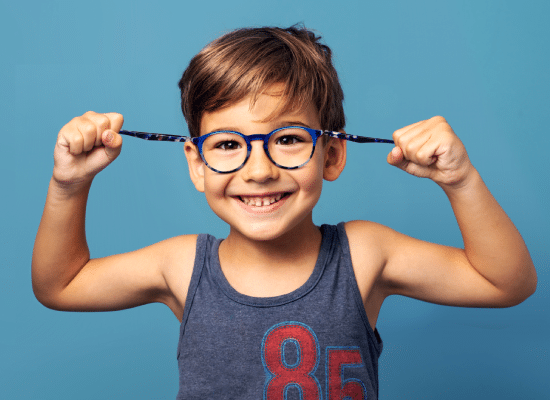 A smiling boy wearing blue glasses, holding the arms of his glasses apart, against a blue background.