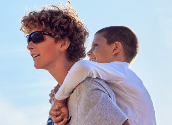 Jeune garçon portant des lunettes de soleil Oakley noires, transportant un enfant sur son dos.