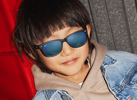 Enfant souriant portant des lunettes de soleil Ray-Ban noires avec un blouson en jean et un sweat à capuche.