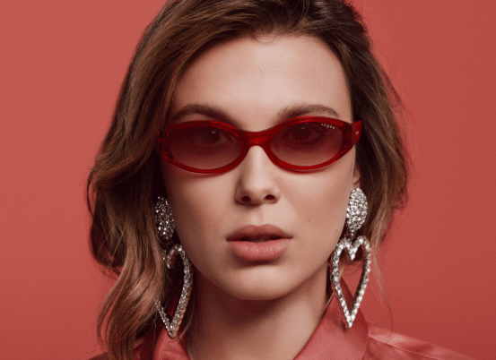 Femme portant des lunettes de soleil rouges, des boucles d'oreilles en forme de cœur et une chemise rose, posant devant un fond rose.