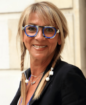 A smiling Sabine B. wearing blue-rimmed rectangular glasses and braids, posing outdoors.