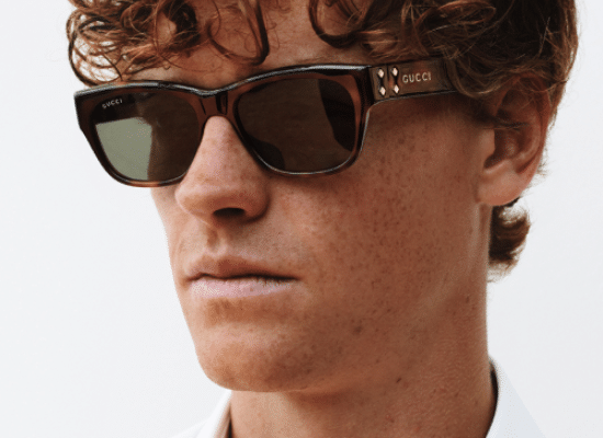 Man with curly hair wearing Gucci sunglasses with thick brown frames and gold temples.