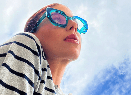 Woman wearing blue sunglasses with thick Kirk & Kirk frames, looking skywards.