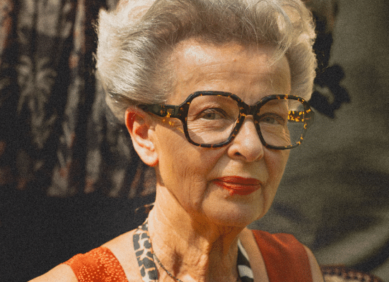 Elderly woman with grey coiffed hair, wearing Moken tortoiseshell glasses, subtle smile.