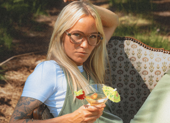 Blonde woman wearing Moken clear-rimmed glasses, sitting in an armchair with a cocktail.