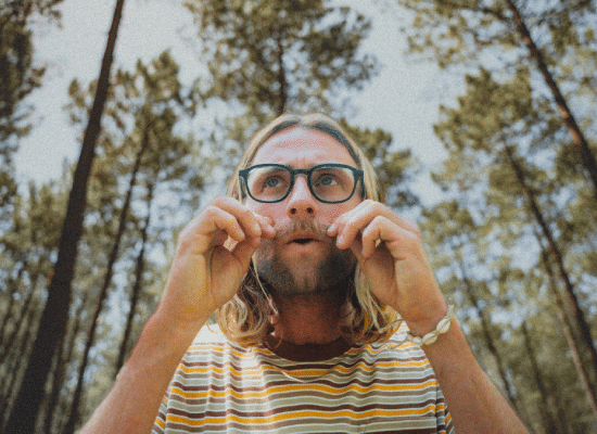 Homme barbu avec des cheveux longs, portant des lunettes de vue Moken à monture verte, debout dans une forêt.