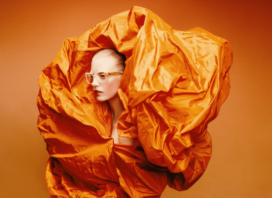 Woman wearing light-rimmed glasses, wrapped in a voluminous orange fabric on a matching background.