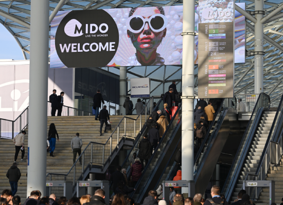 Entrée d'un événement avec une bannière numérique affichant "MIDO WELCOME" et des visiteurs montant les escaliers mécaniques vers l'exposition.